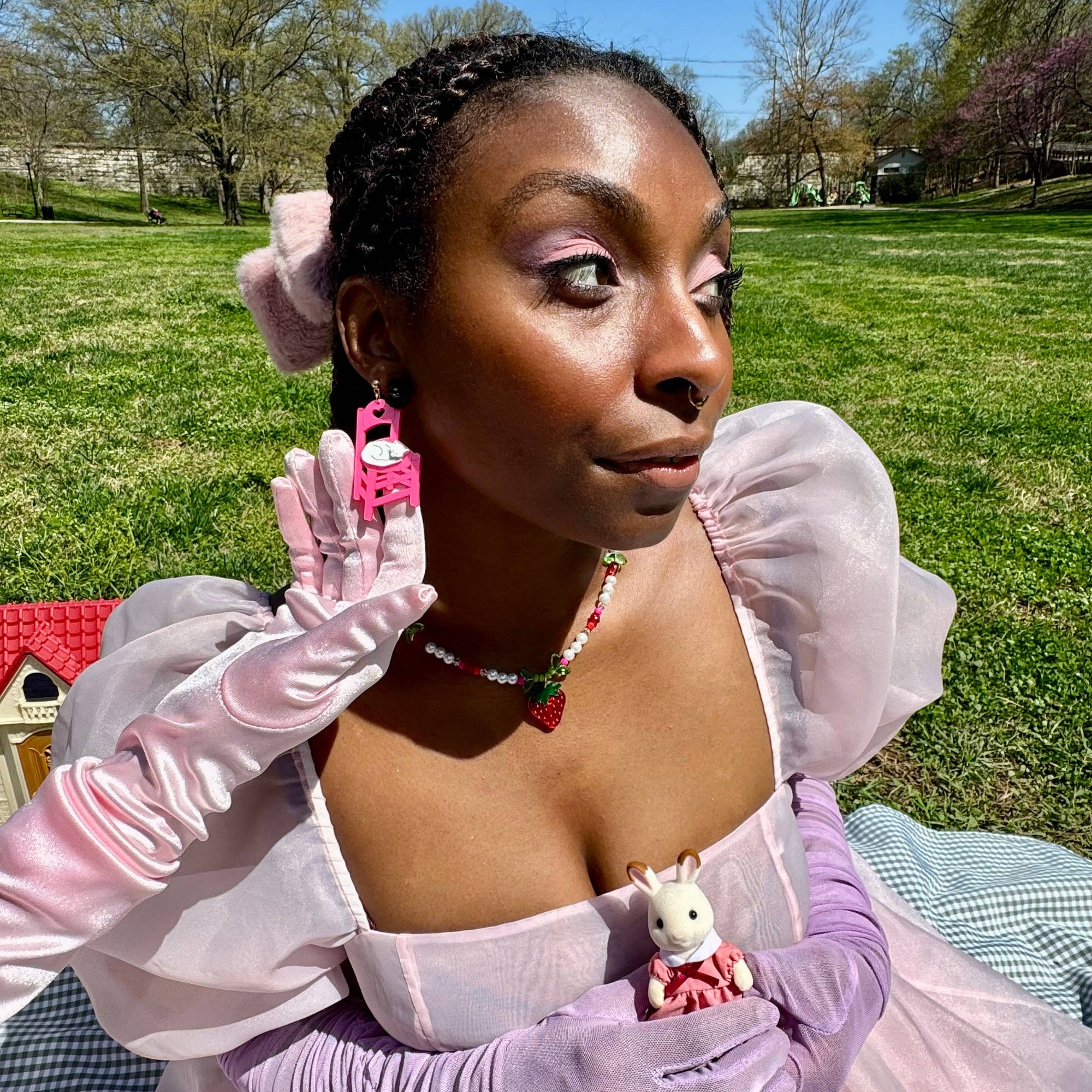 Person in a pink dress with puffed sleeves and gloves, holding a small rabbit figure, outdoors.