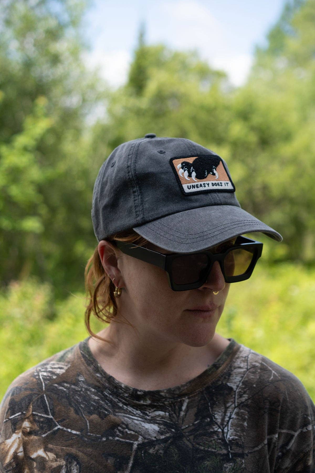 Person wearing a cap with a patch and sunglasses, standing in a natural setting.
