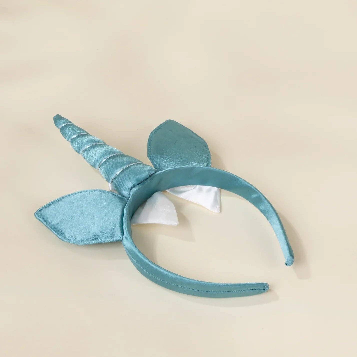 A light blue satin unicorn headband with a horn and ears, featuring a small white bow, displayed on a neutral beige background.