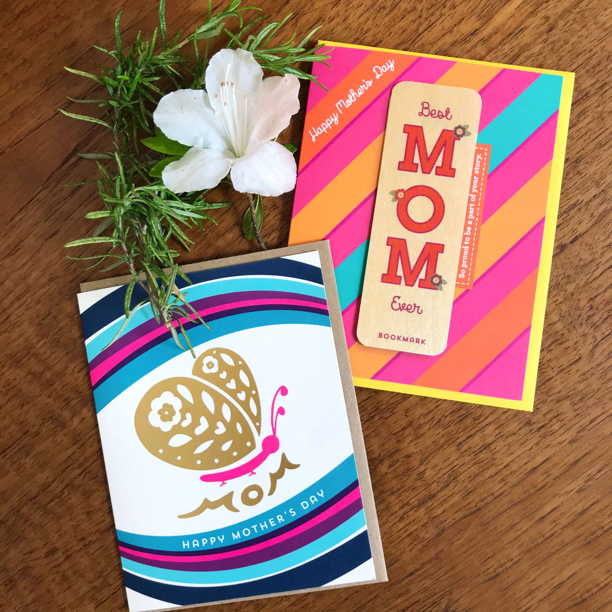 Two Mother's Day cards on a wooden surface with a plant and flower.