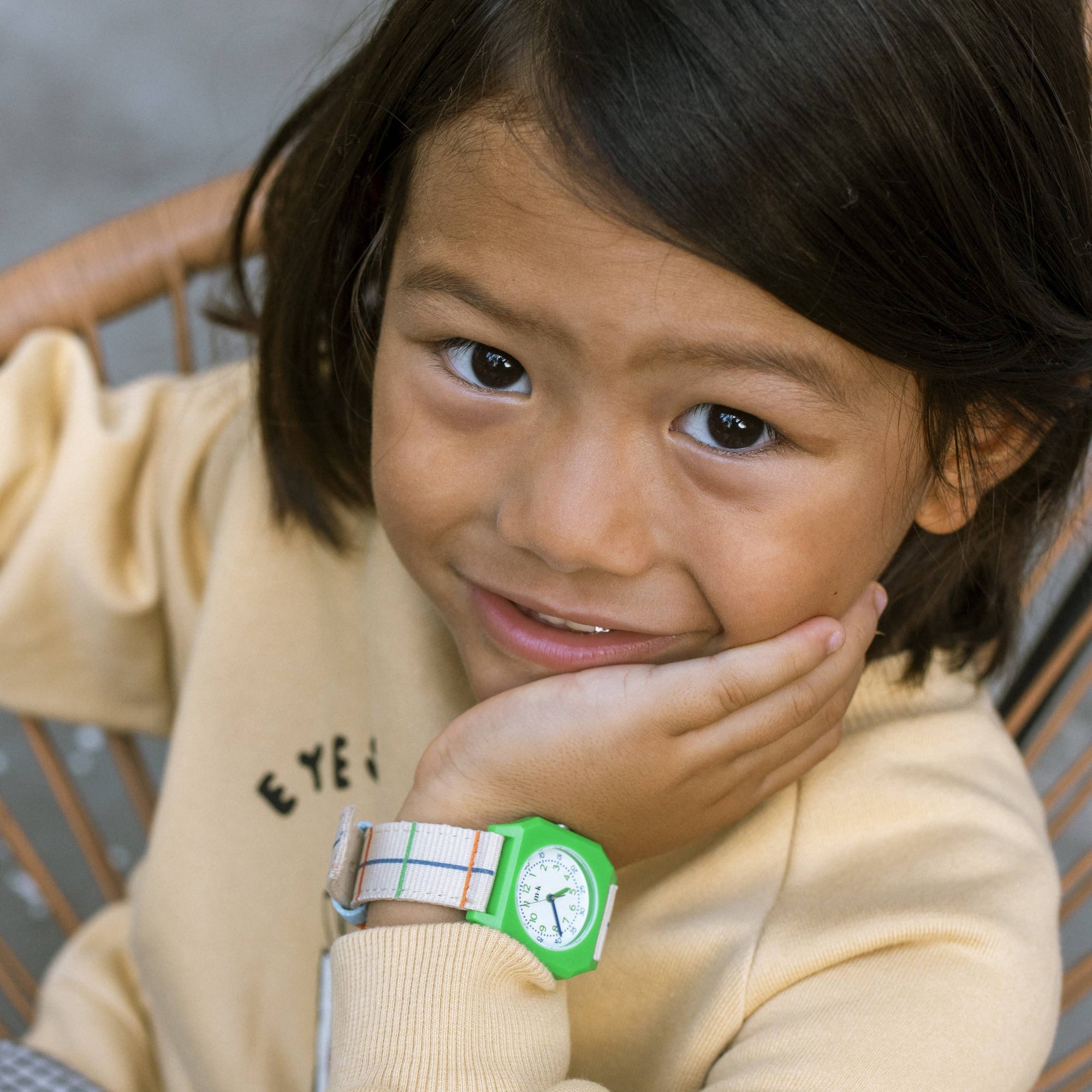 Child wearing a beige sweatshirt with text, holding a green watch, sitting on a wooden chair.