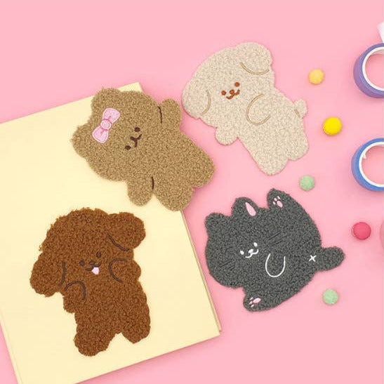 Three textured plush dog-shaped stickers or patches in brown, beige, and dark gray, arranged on a pink background with a cream card and small colorful pom-pom accents.