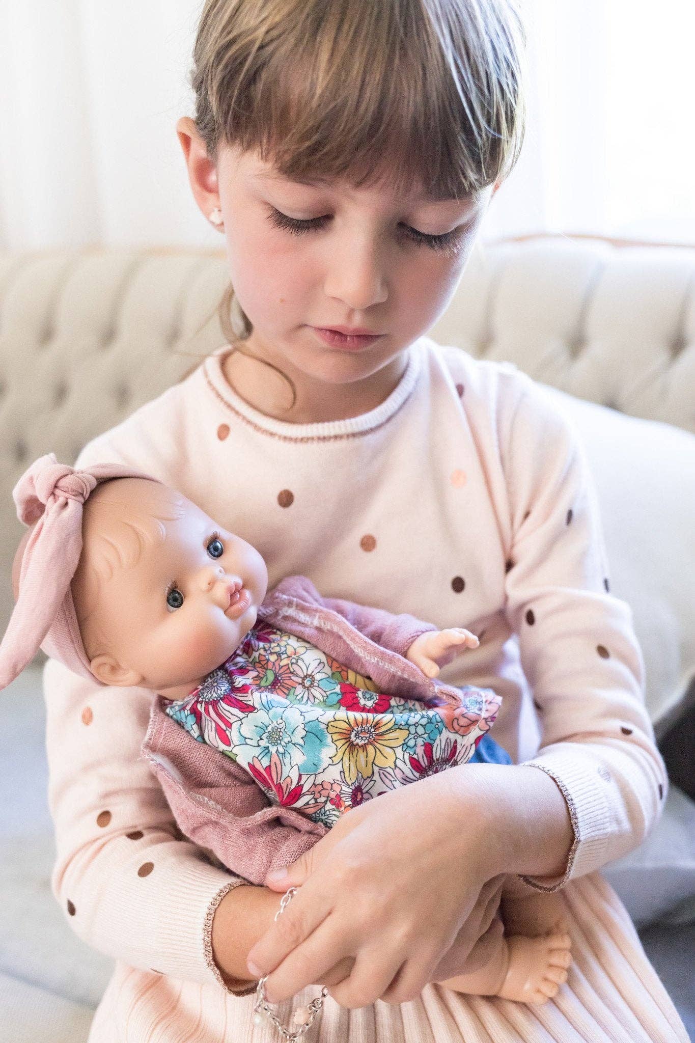 Child holding a baby doll with a floral blanket on a light-colored couch.