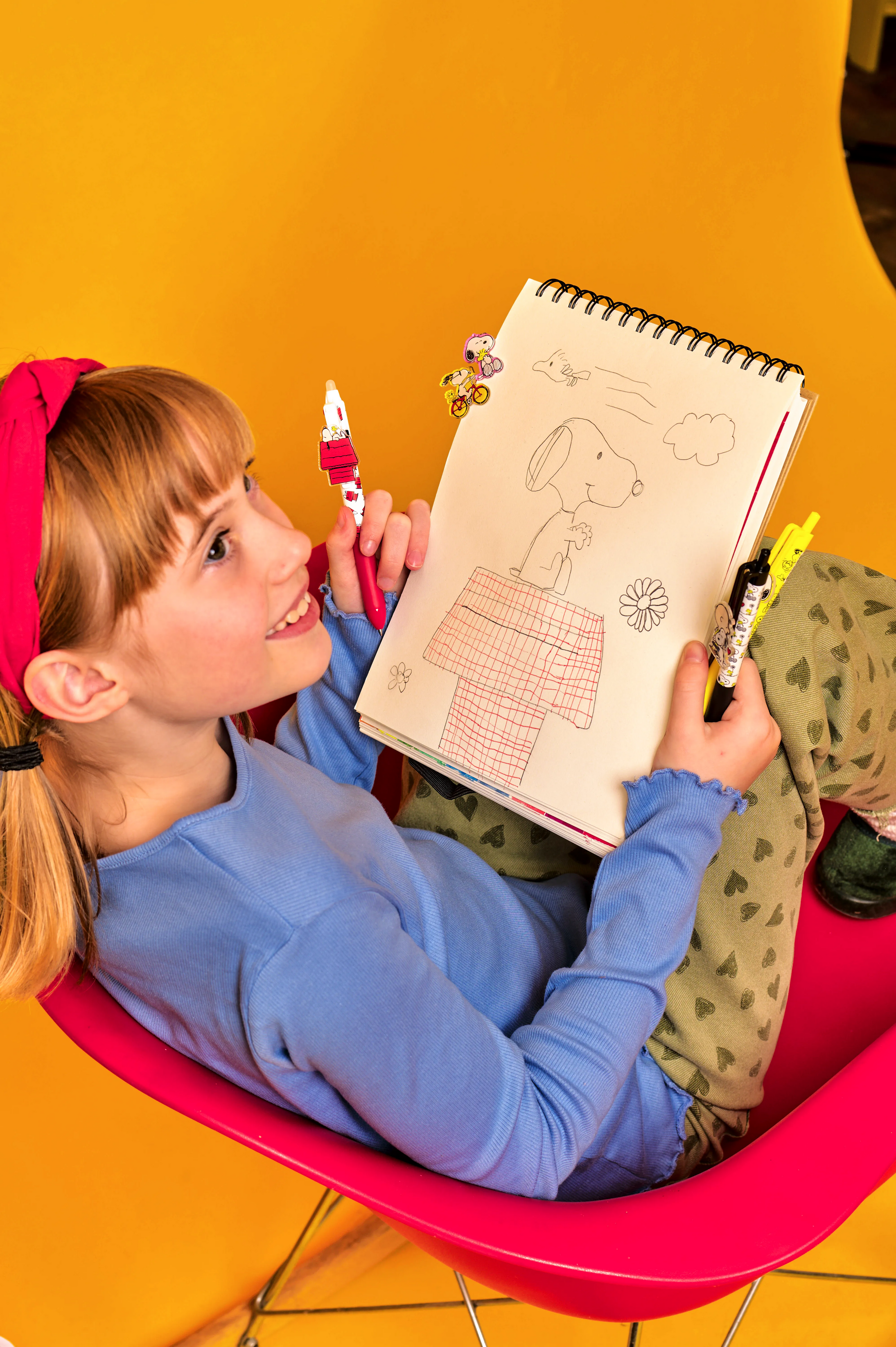 Child sitting on a pink chair holding a drawing book with illustrations against a yellow background