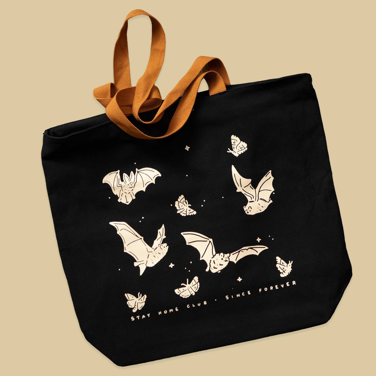 A black canvas tote bag with brown fabric handles, decorated with cream-colored bat and butterfly illustrations and the text 'stay home club - since forever' on a solid beige background.