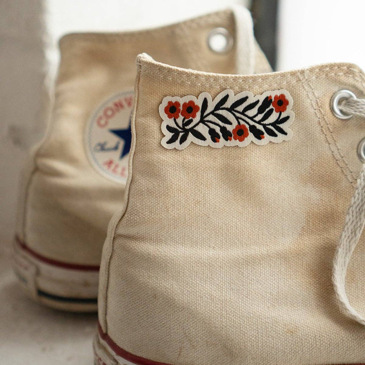 A close-up of a beige canvas high-top sneaker with a decorative embroidered floral patch on the side, alongside another similar sneaker in the background.