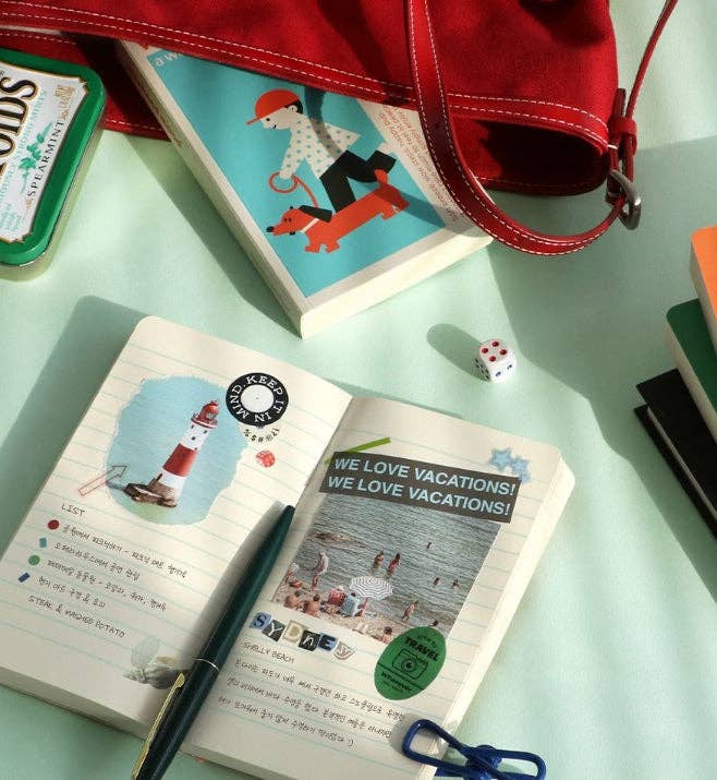 Open notebook with travel-themed pages on a light green surface, with a red bag and other items in the background.