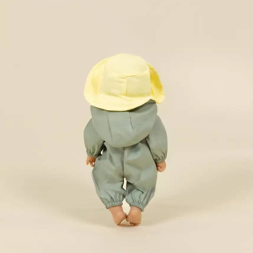 A doll viewed from behind wearing a pale sage green hooded jumpsuit and a soft yellow bucket hat, standing against a plain warm beige studio background.