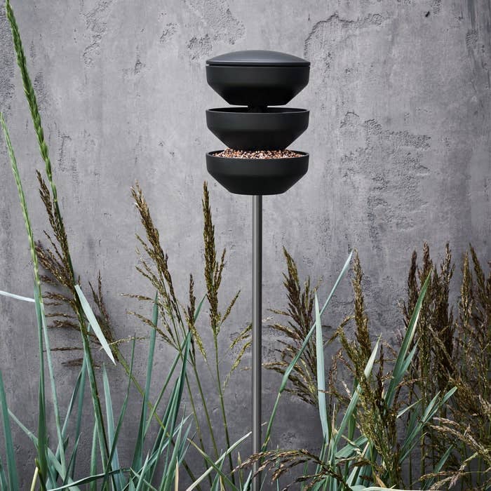 Black tiered bird feeder against a concrete wall with grasses in the foreground