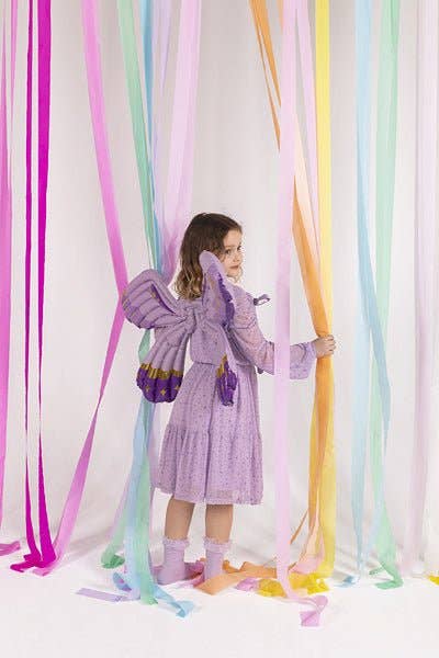 A young girl stands in a studio wearing a lavender-purple fairy costume with large wings, holding a long pastel ribbon. Multiple colorful hanging ribbons in pink, blue, green, yellow, and orange are suspended against a white background.