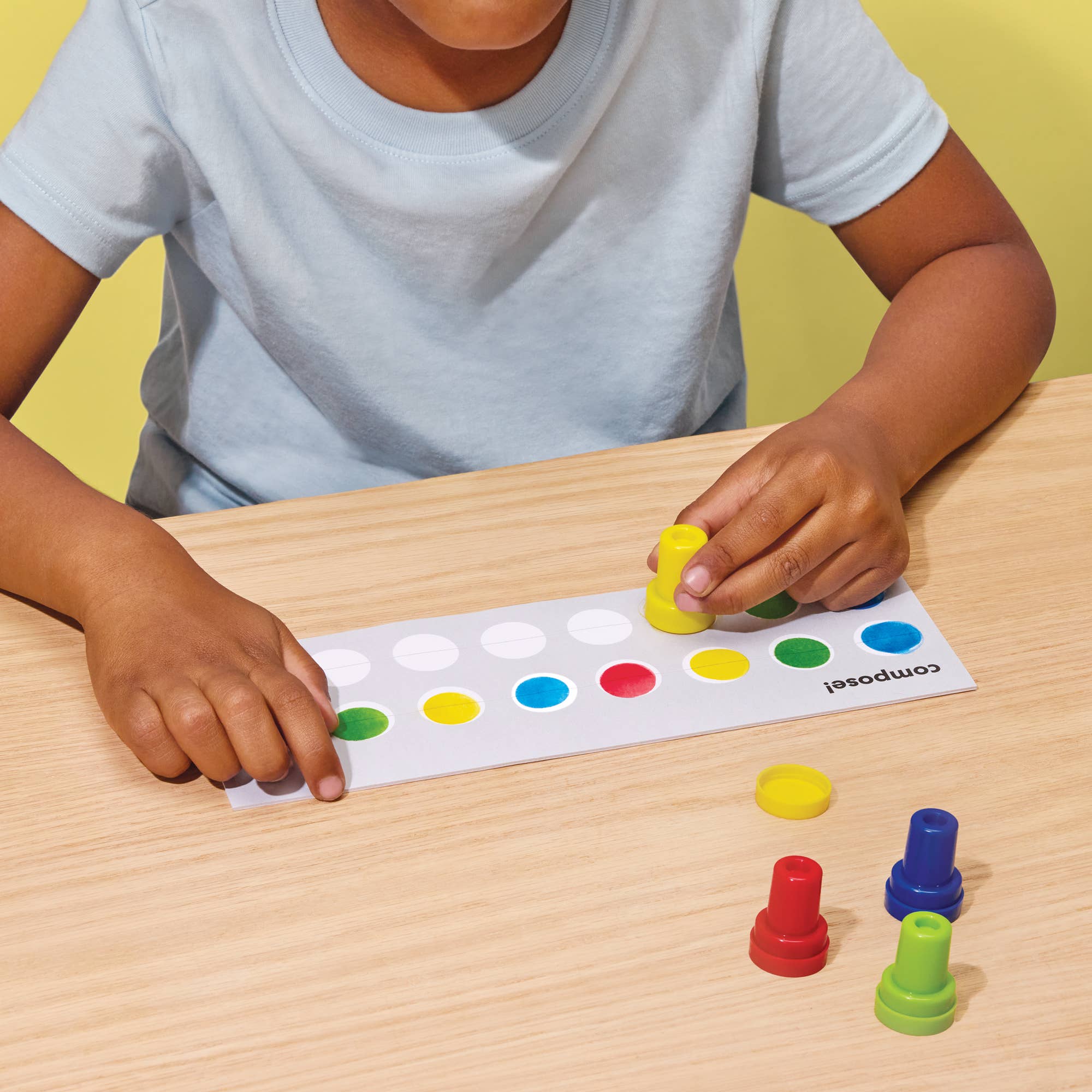 A child wearing a light blue t-shirt is shown from the chest down, playing with a white composition board that has colored circular spots and small colorful pegs on a light wood table.