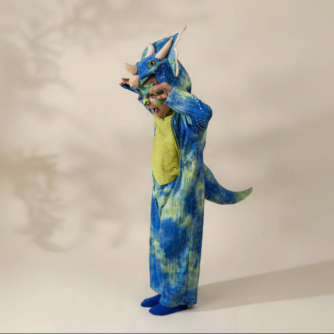 A child wearing a blue and green dinosaur costume with a horned headpiece, standing against a plain light beige studio background.