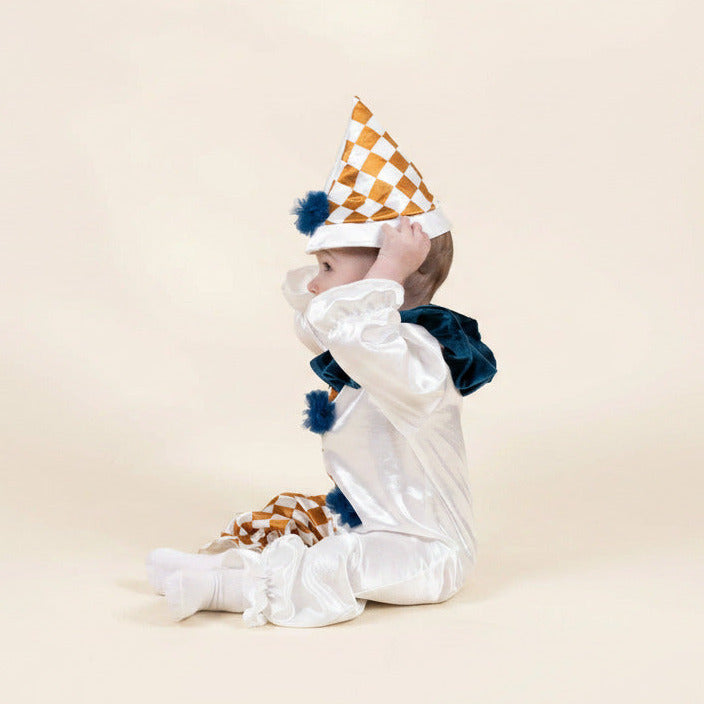 A baby wearing a white clown costume with a gold and white checkered party hat, blue pom-poms, and a dark blue ruffled collar, sitting against a solid beige background.
