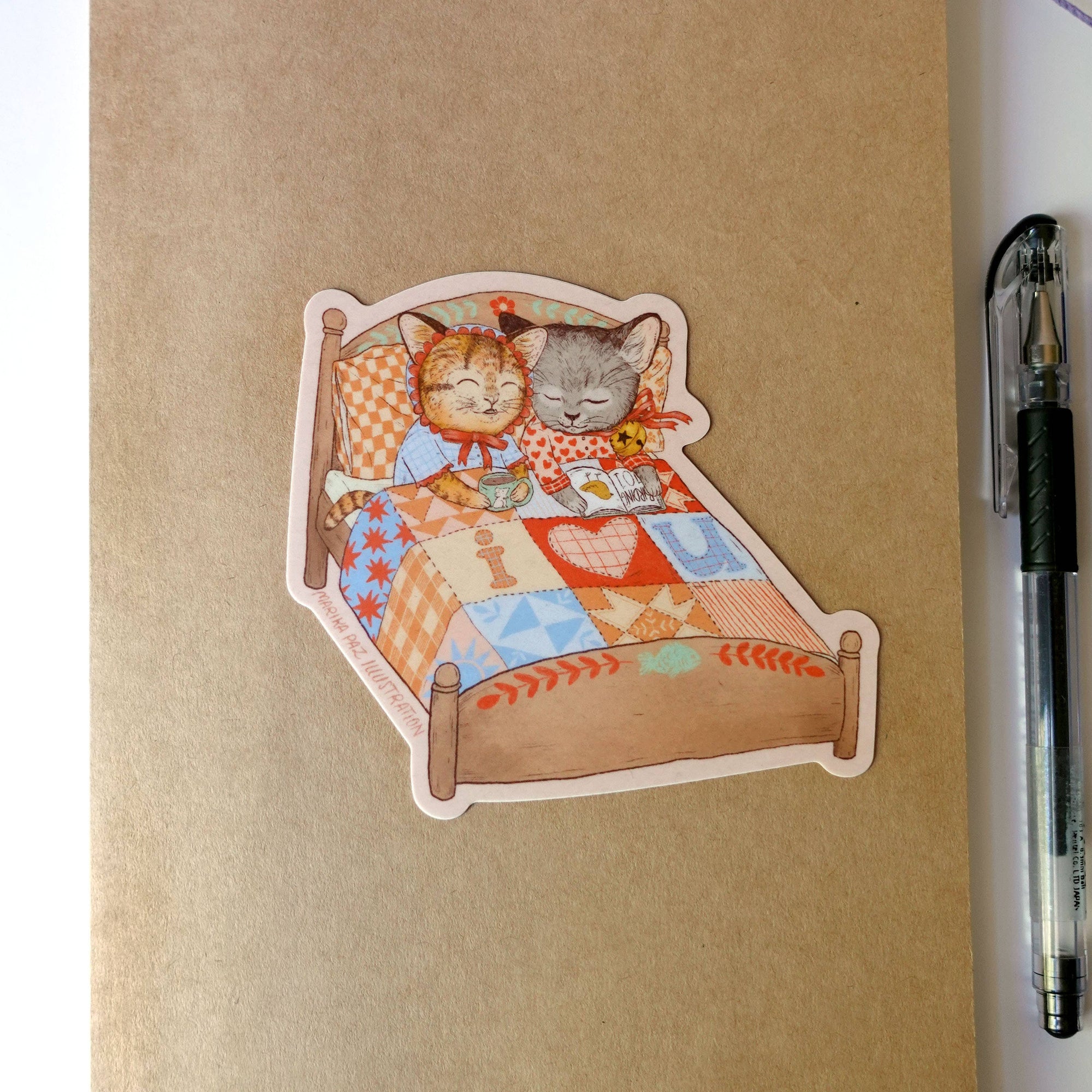 Notebook with a sticker of two cats on a bed, next to a pen and yarn.