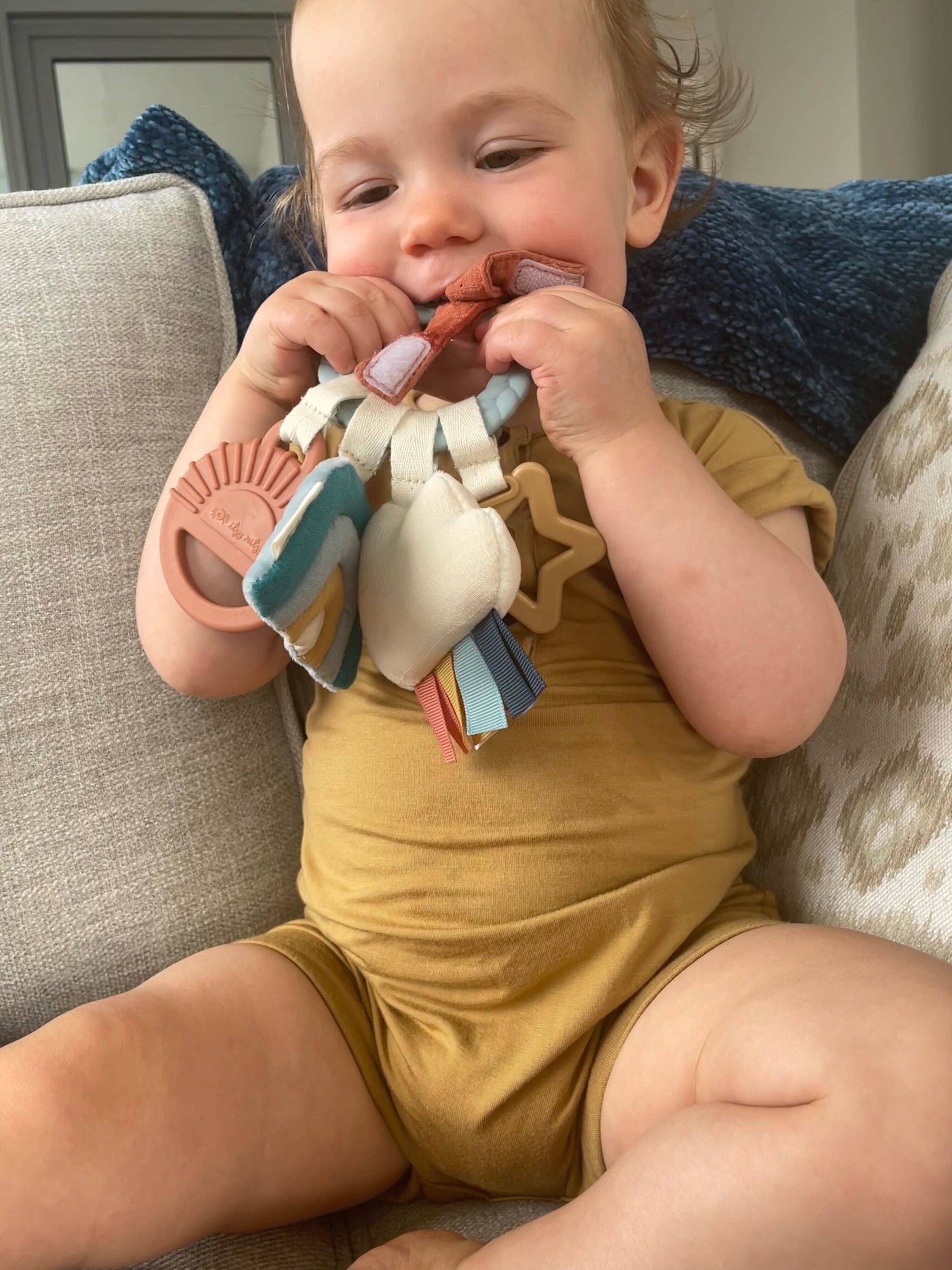 Baby in a yellow outfit playing with a colorful teething toy on a couch.