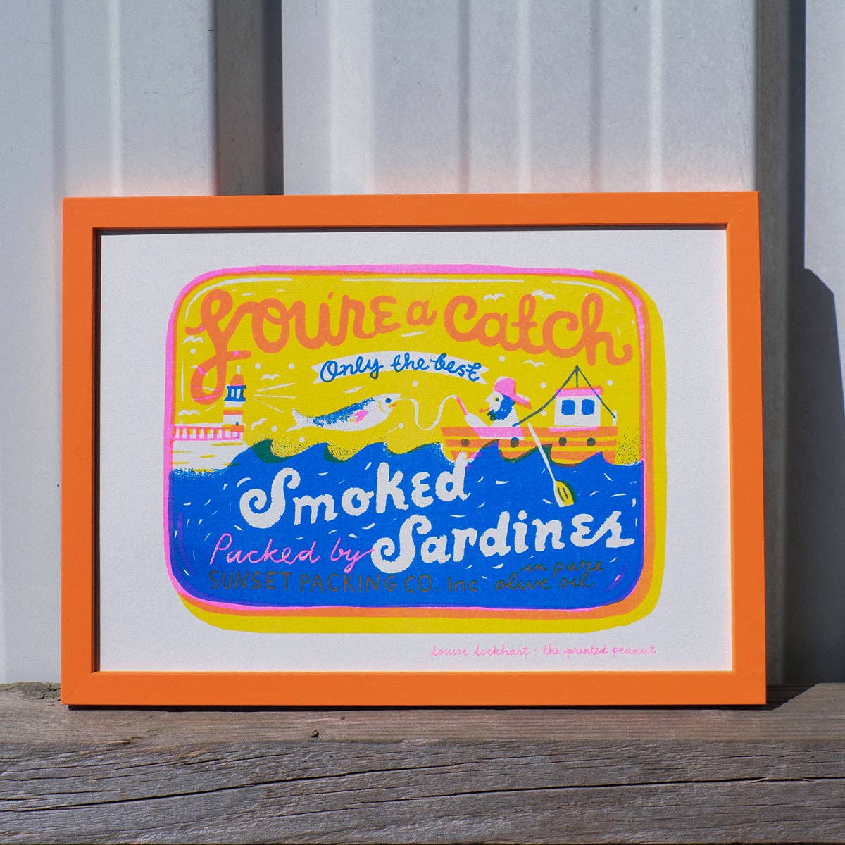Framed artwork with a colorful design of a can of sardines and text on a wooden surface.