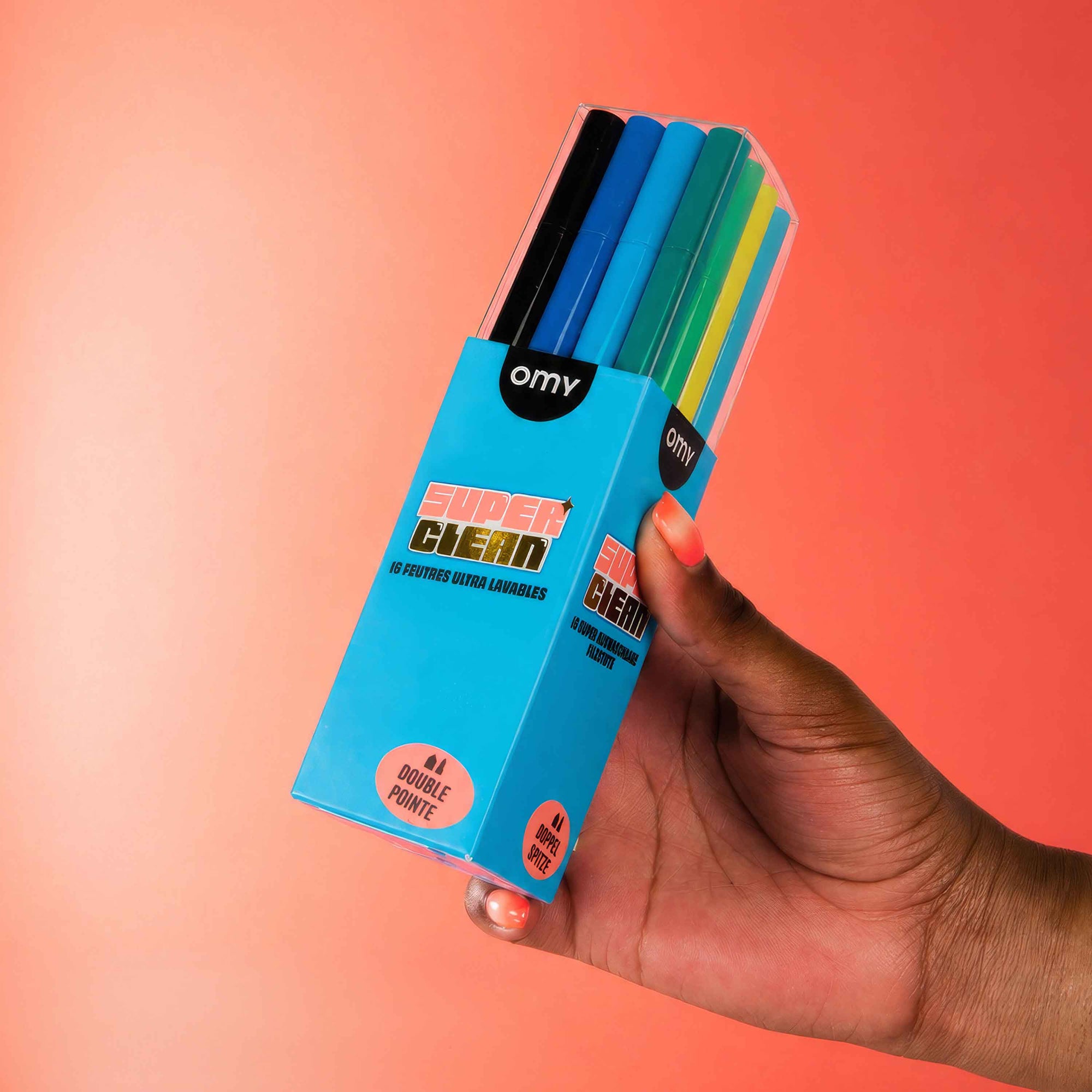 A hand holding a box of OMY Super Clean 12-Count Double Point Marker pens, showing the colorful tips of the markers through a clear plastic window on the box, set against a solid orange background.