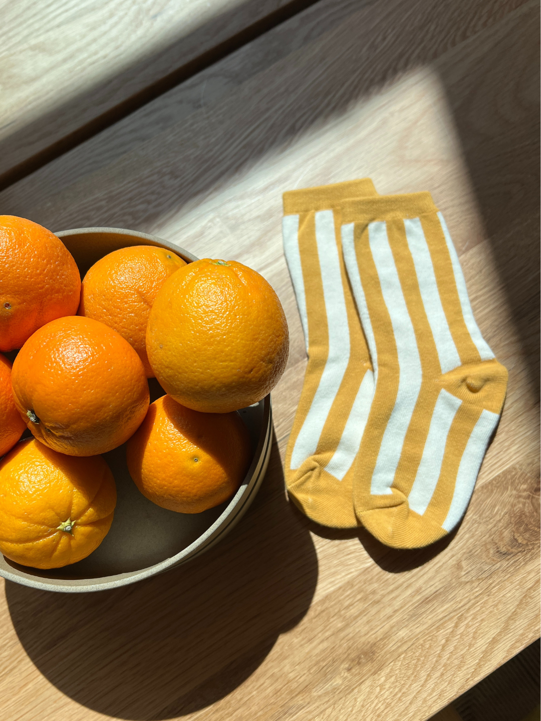 Bowl of oranges next to a pair of mustard yellow socks with white stripes on a wooden surface.