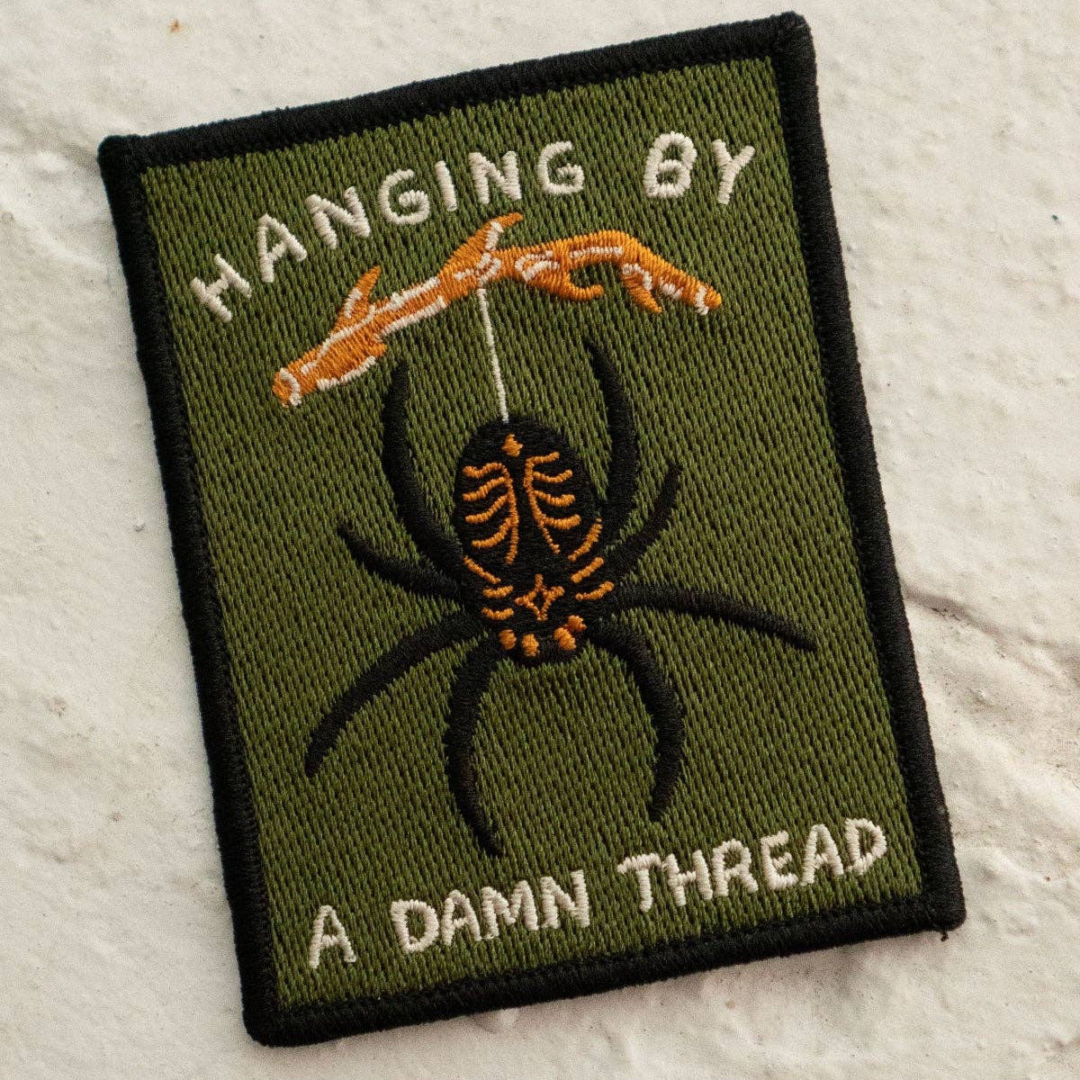 Patch with spider design and text 'Hanging by a Damn Thread' on a textured surface