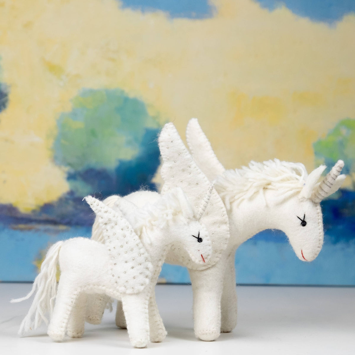 Two white unicorn toys in front of a colorful landscape painting.