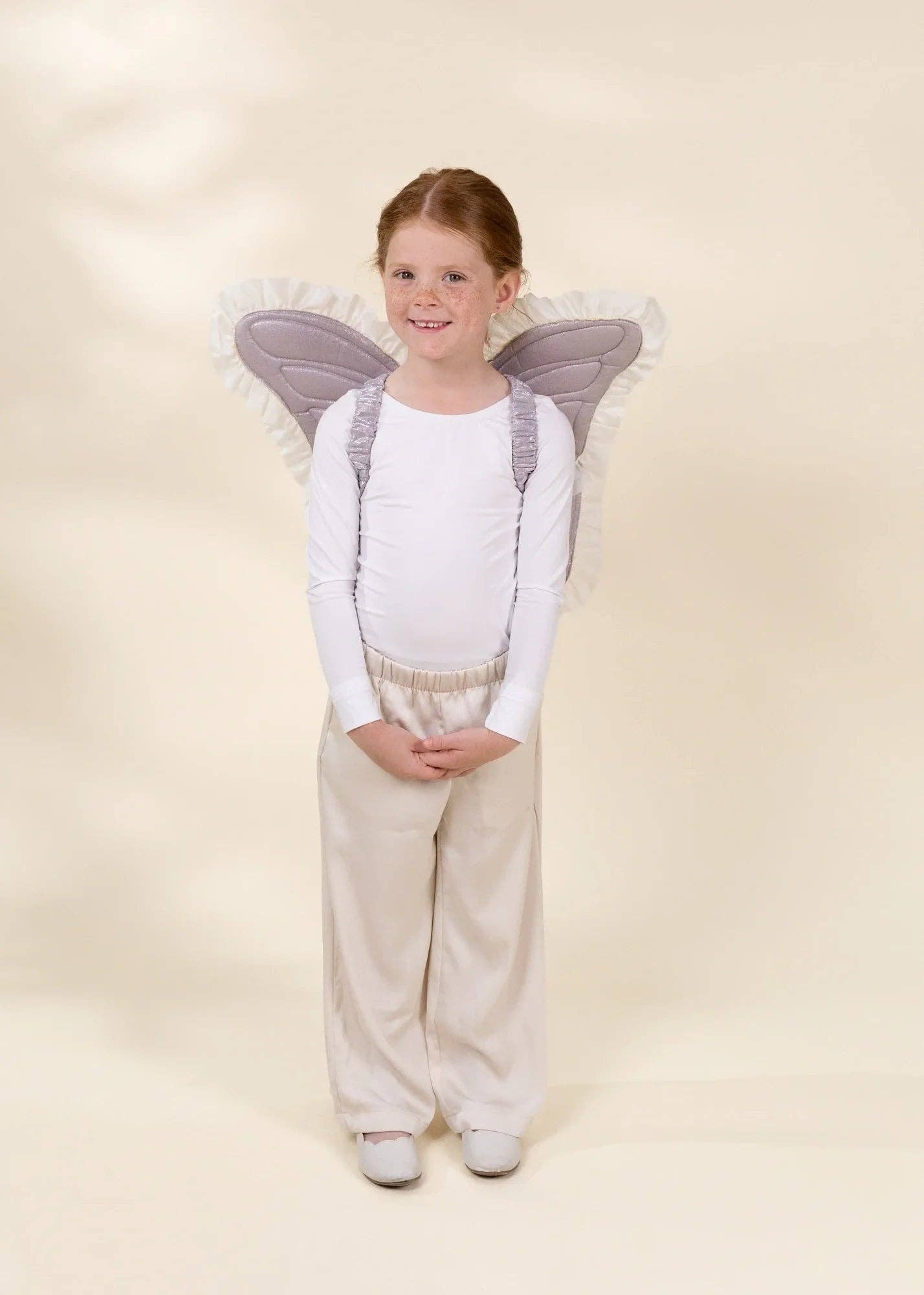 A young girl with red hair stands wearing a white long-sleeve top, cream pants, and gray angel wings with white tulle trim, photographed against a plain warm cream studio background.