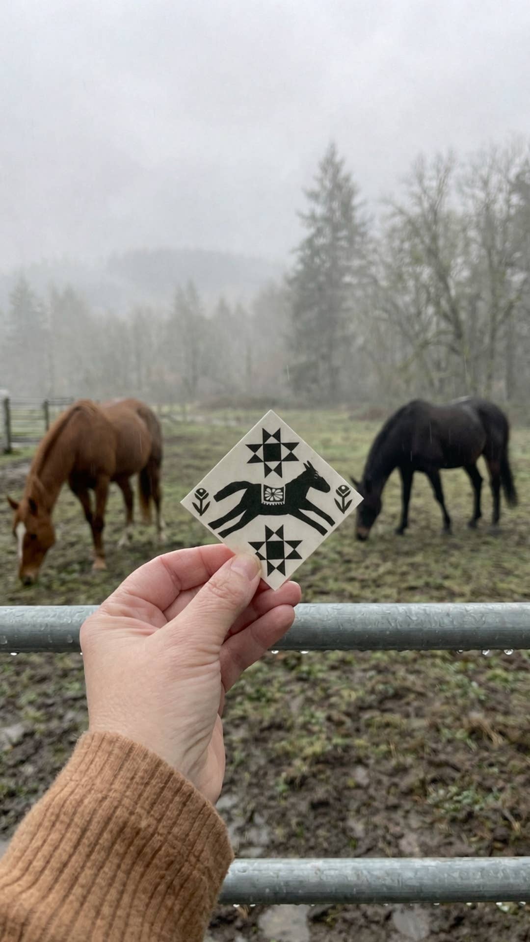 Hand holding a tile with a black and white design of a horse in front of two horses grazing in a misty field.