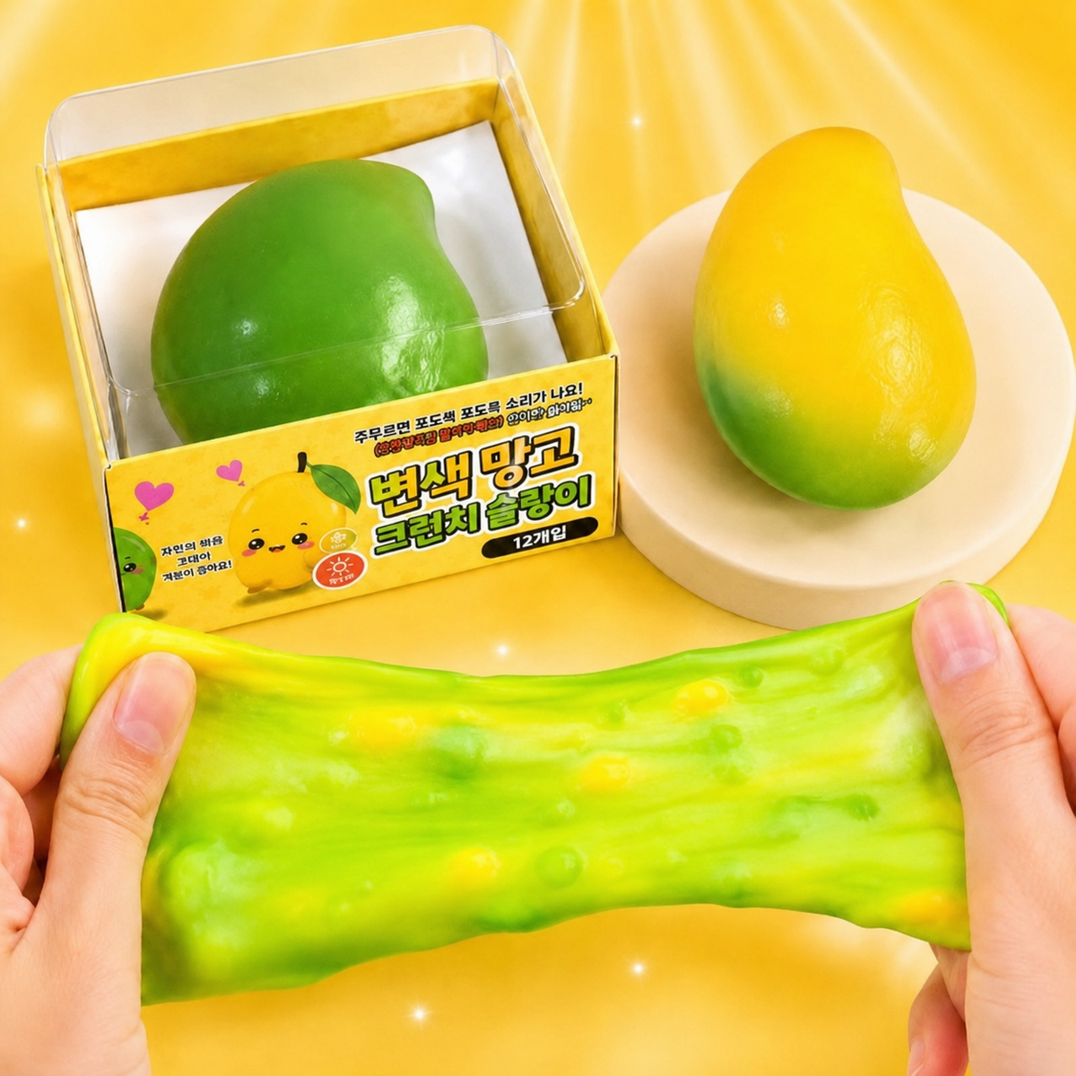 A green and yellow squishy toy shaped like a mango, shown being stretched by two hands in front of its yellow retail box, with another mango-shaped piece on a small white dish against a bright yellow background.