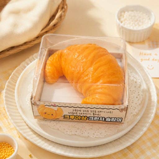 A croissant-shaped novelty food item in a clear plastic box, displayed on a white plate with a yellow checkered cloth and small bowls in the background.