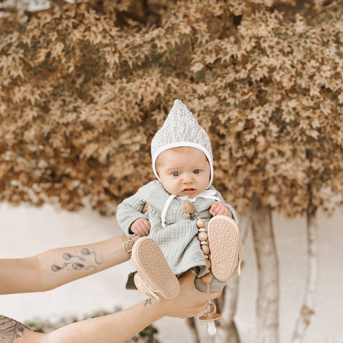 A baby wearing a light gray knit bonnet and matching outfit is held up by tattooed arms in front of a blurred tree with brown leaves.