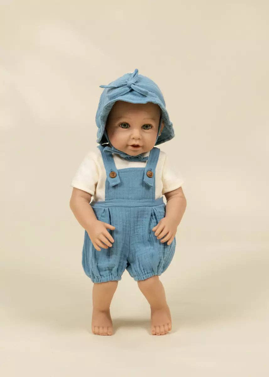 A baby doll or toy dressed in light blue denim-style overalls with a matching bonnet, a white short-sleeve shirt, and bare feet, standing against a plain warm beige studio background.