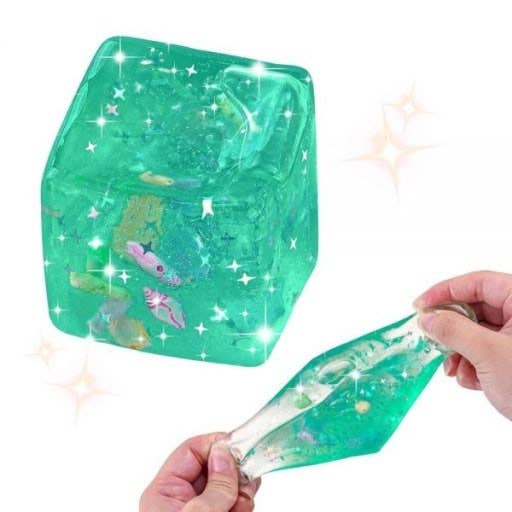 A translucent green slime or gel cube with embedded glitter and small fish-like shapes, shown alongside hands stretching a matching translucent green gel strip on a white background with sparkles.