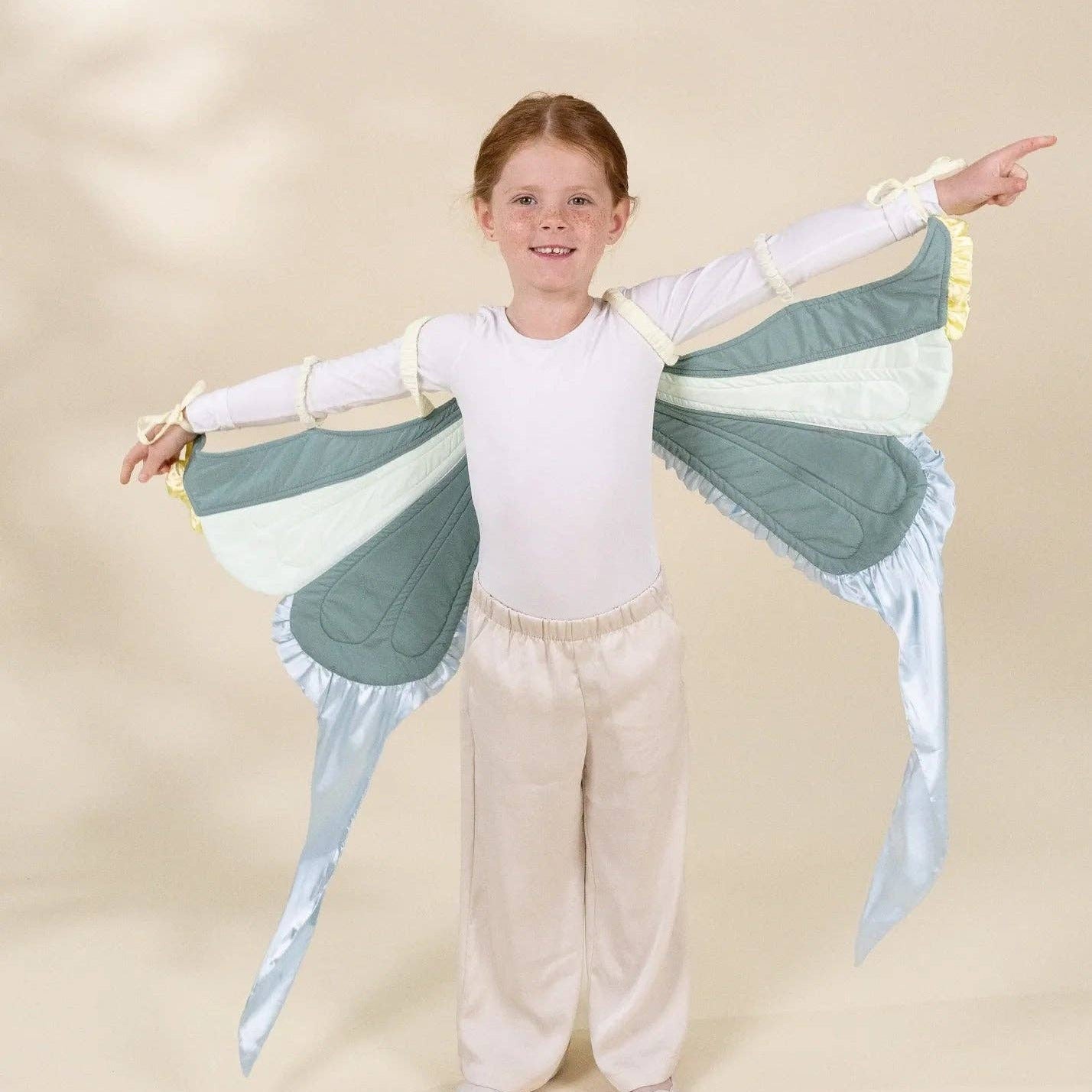A young girl with light brown hair stands in a studio wearing a butterfly wing costume over a white long-sleeve top and cream pants. The wings are light blue and mint green with pale yellow trim and long flowing tails.