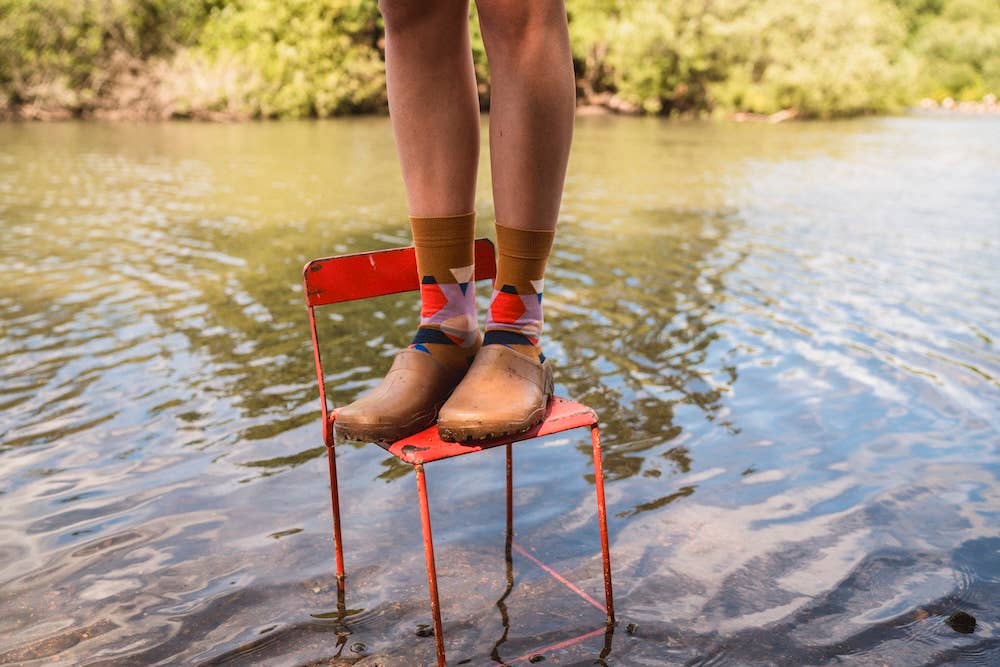 Person standing on a red chair in water with feet wearing brown boots and colorful socks.