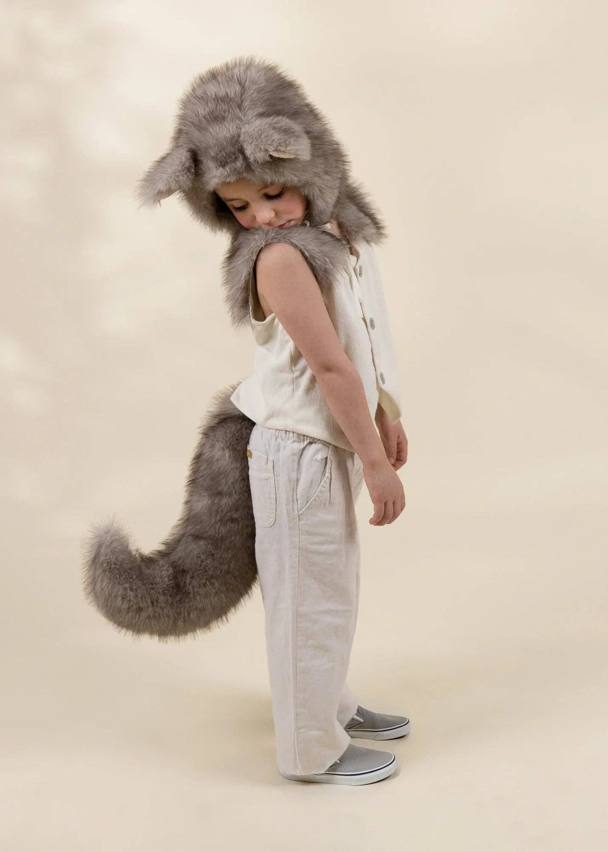 A child wearing a gray faux-fur wolf costume hat with a long tail, a cream sleeveless vest, and cream pants, standing against a warm beige studio background.
