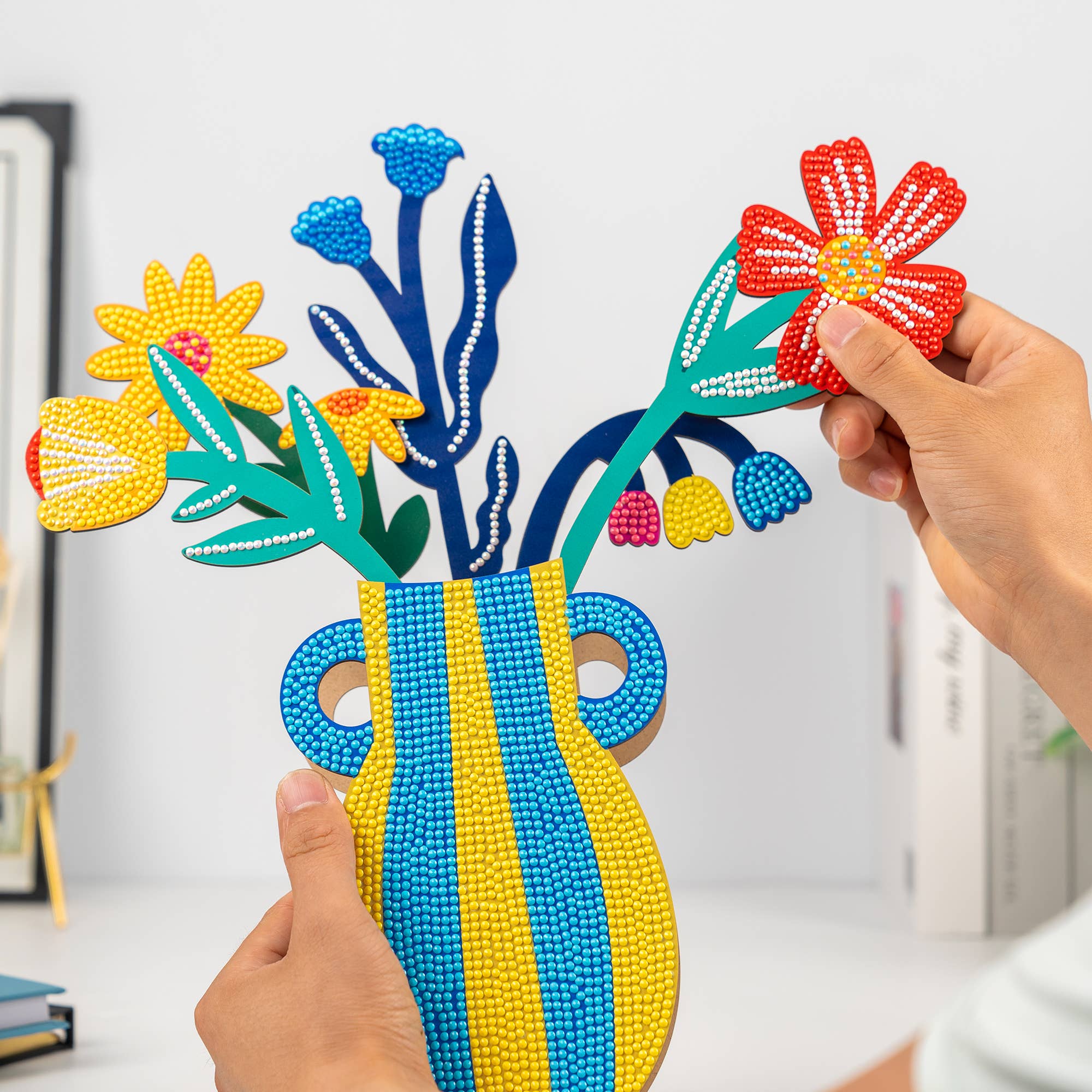 Colorful beaded flower arrangement and vase held by hands against a white background