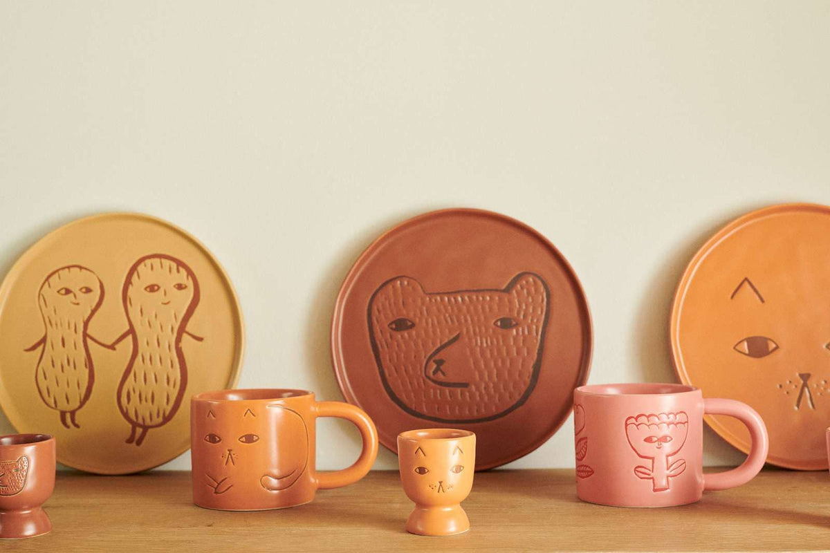 A collection of handcrafted ceramic tableware in terracotta, orange, and pink, featuring minimalist cartoon-style drawings of peanuts, a bear, and a face on plates and mugs, displayed on a wooden surface against a light beige wall.