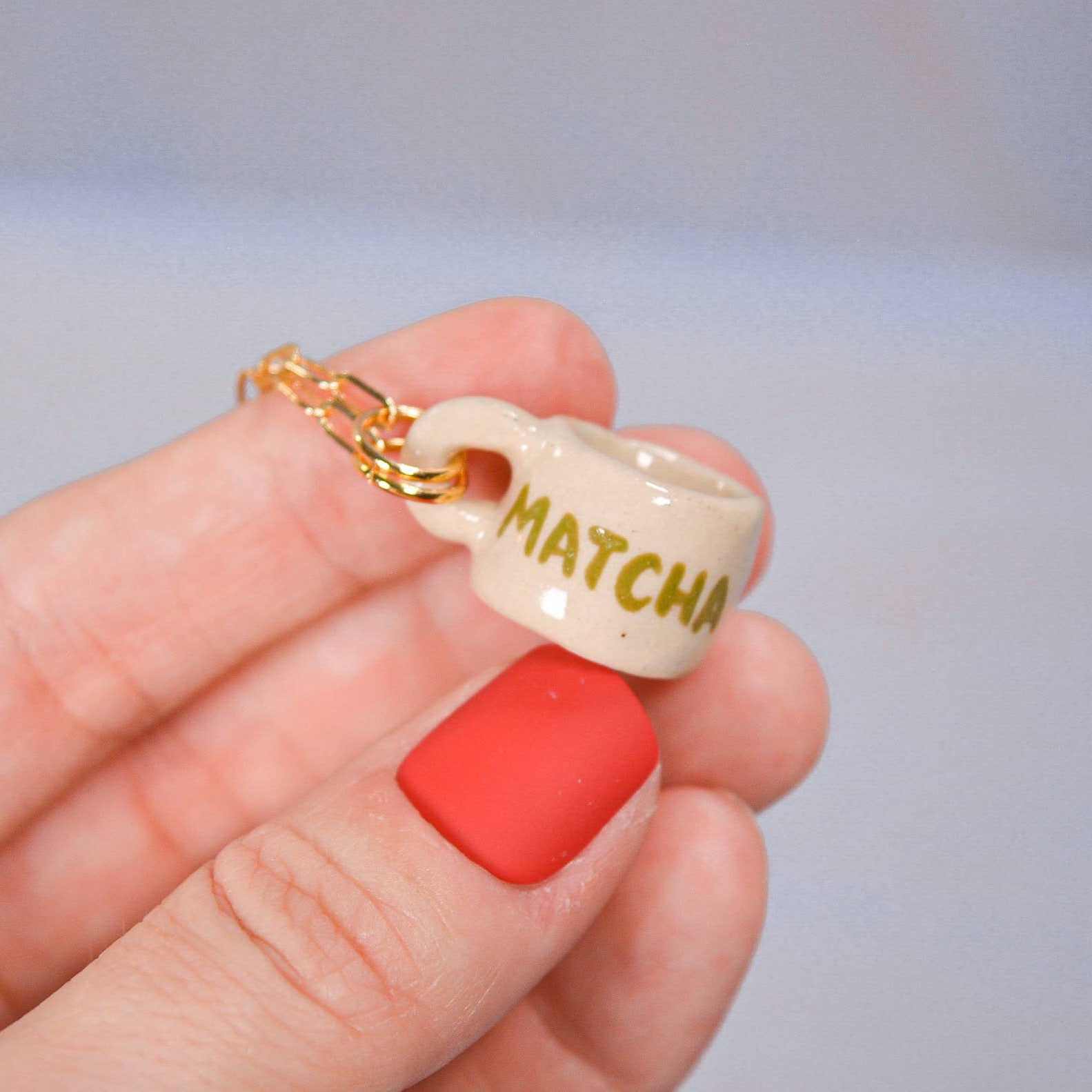 Small ceramic cup charm with 'MATCHA' text held in a hand against a neutral background
