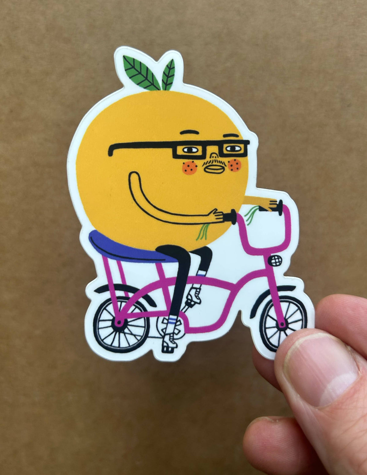 A die-cut sticker featuring a cartoon orange character wearing glasses and riding a pink bicycle, held by a person's fingers against a brown cardboard background.