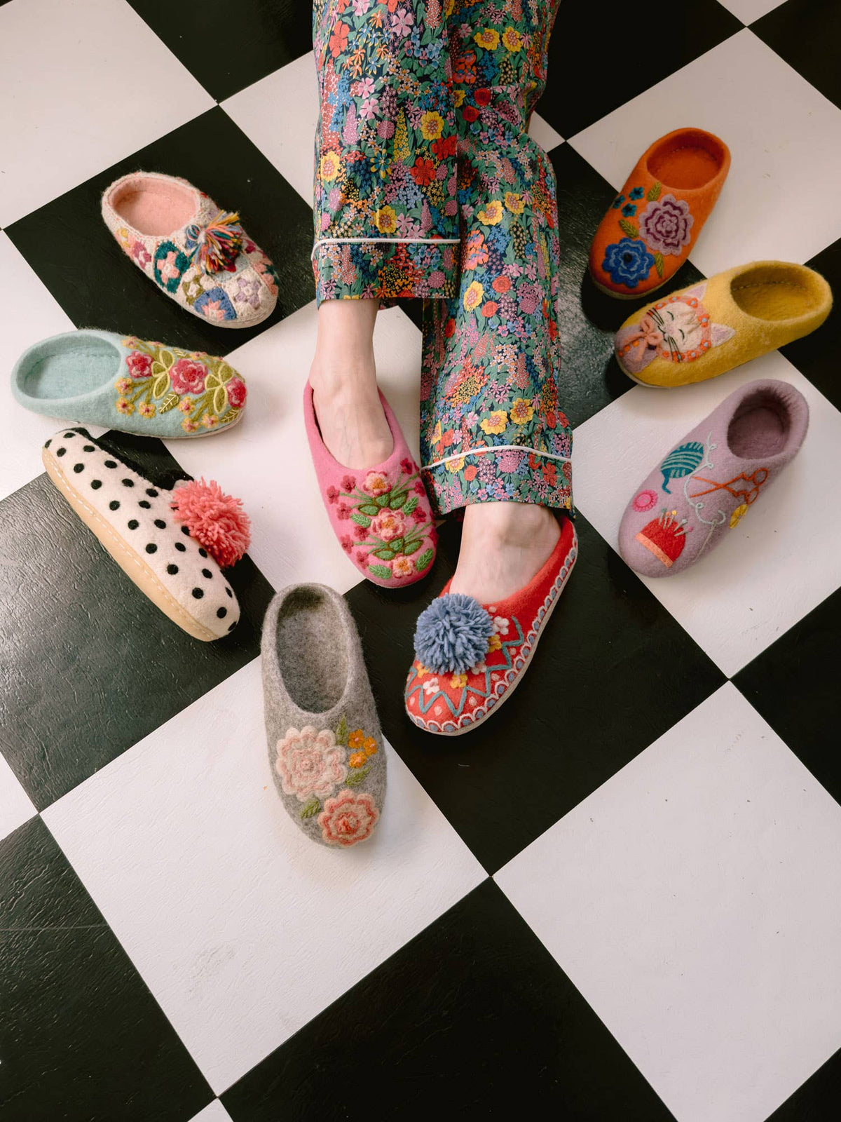 A top-down view of a person wearing colorful floral slip-on clogs and matching floral pants, standing on a black-and-white checkered floor with several other floral slip-on shoes arranged around them.
