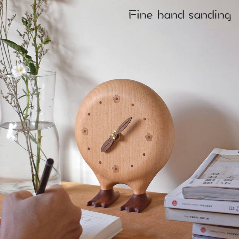 Wooden clock with feet on a table next to books and a vase with plants.
