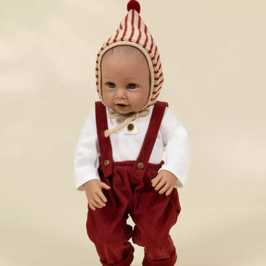 A baby doll wearing a red and cream striped bonnet, a white long-sleeve shirt, and red corduroy overalls, standing against a plain beige background.
