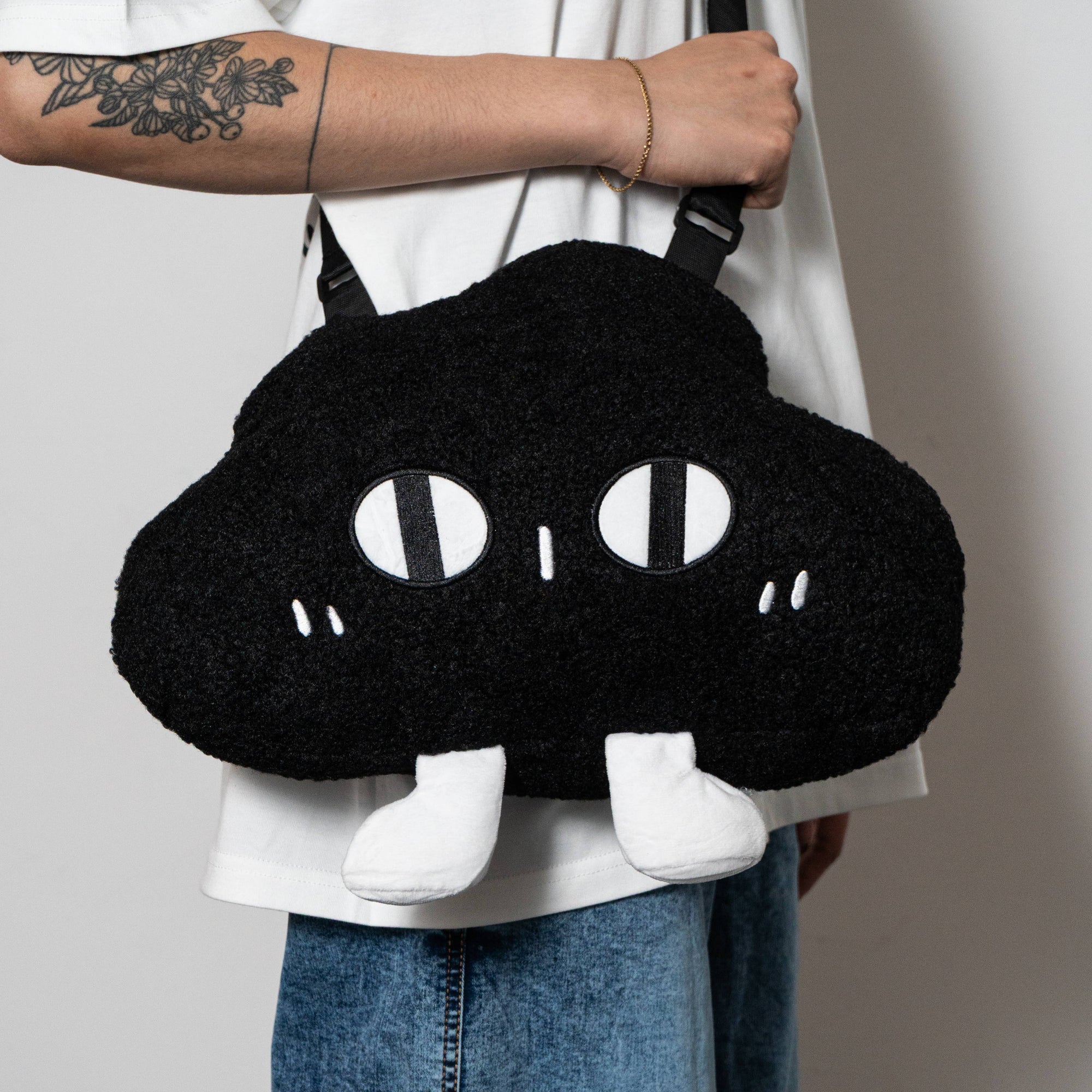 Person holding a black cloud-shaped bag with white eyes and feet against a neutral background
