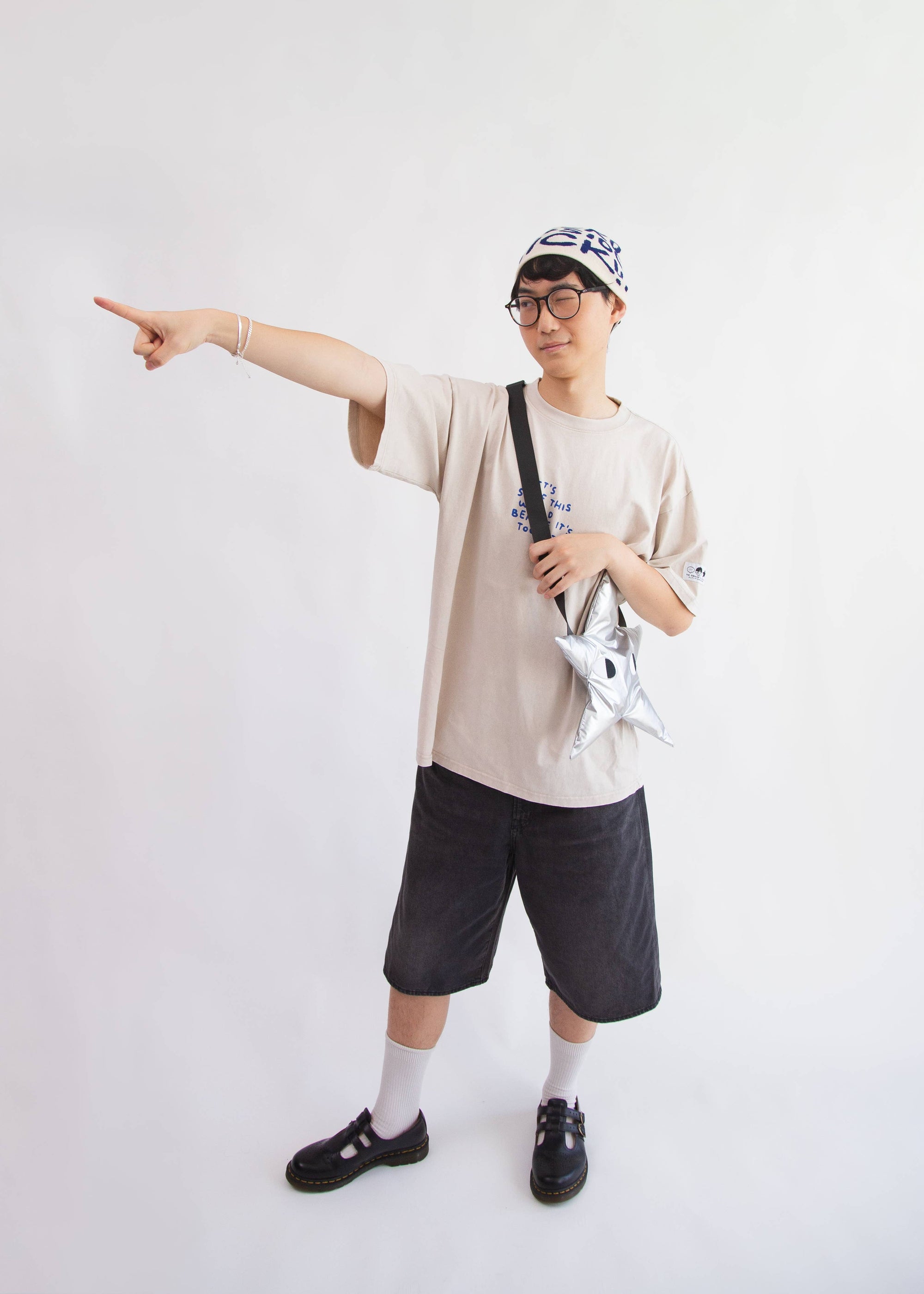 Person wearing a beige t-shirt, black shorts, and a cap, holding a bag and pointing with a white background