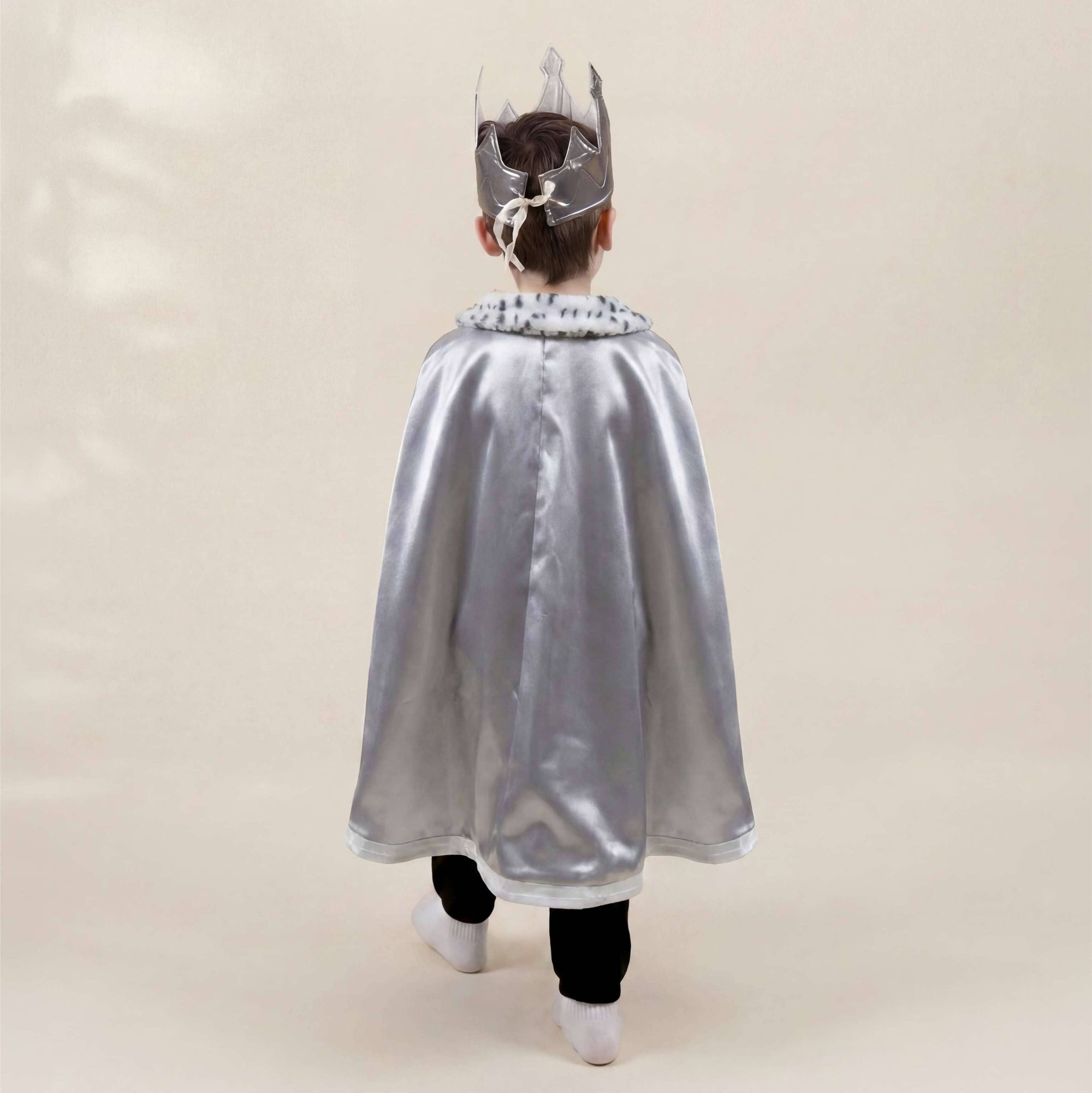 A child viewed from behind wearing a shiny silver cape with a decorative collar and a matching silver crown, standing against a plain warm beige studio background.