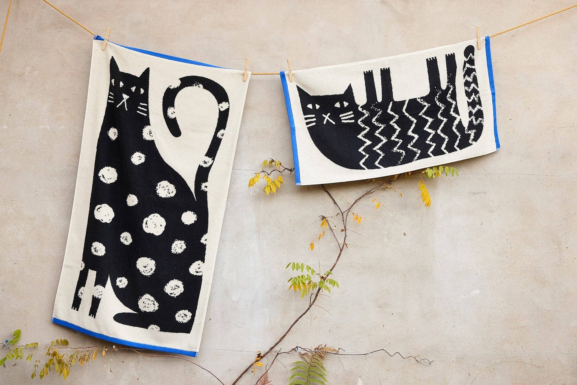 Two towels with cat designs hanging on a line against a neutral background