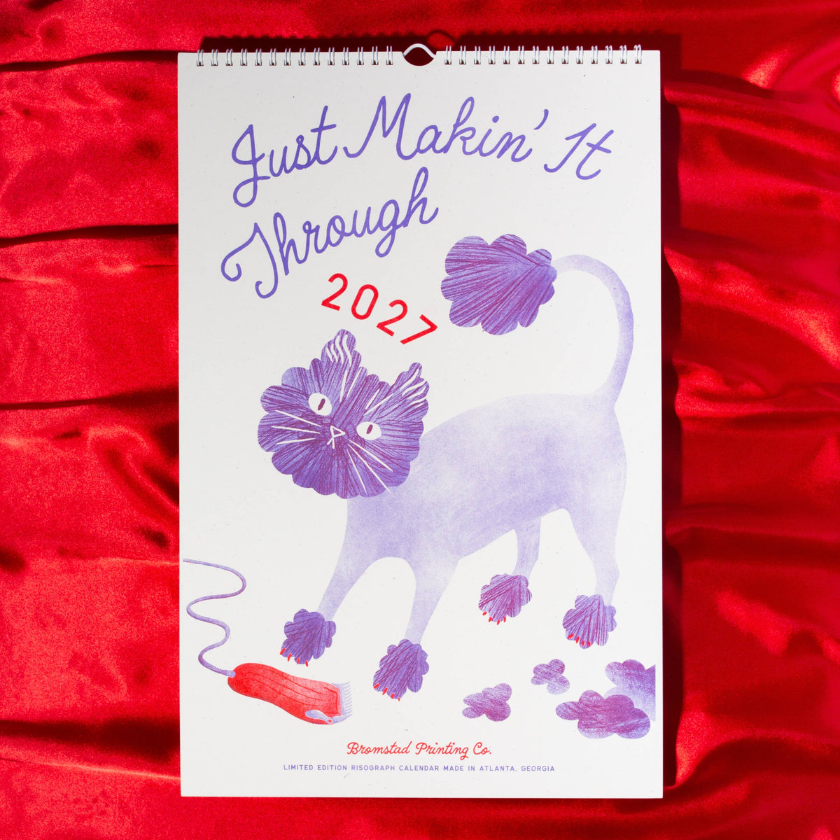 A spiral-bound wall calendar page featuring a purple cat with pom-poms and the text 'Just Makin' It Through 2027,' displayed against a shiny red fabric background.
