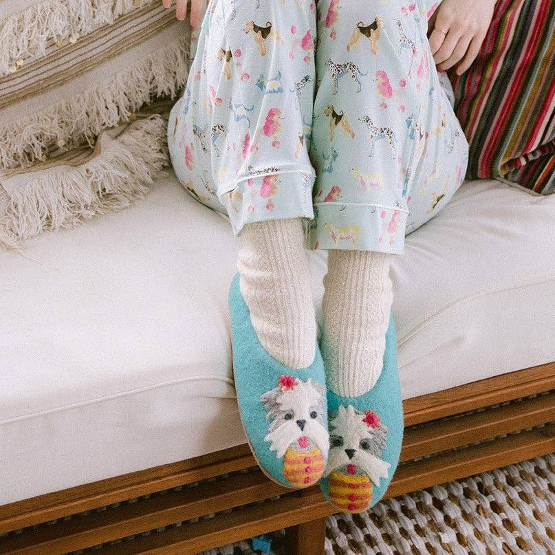 A person sitting on a white sofa wearing pastel pajama pants with a dog print, white ribbed socks, and light blue slip-on slippers featuring  dogs 