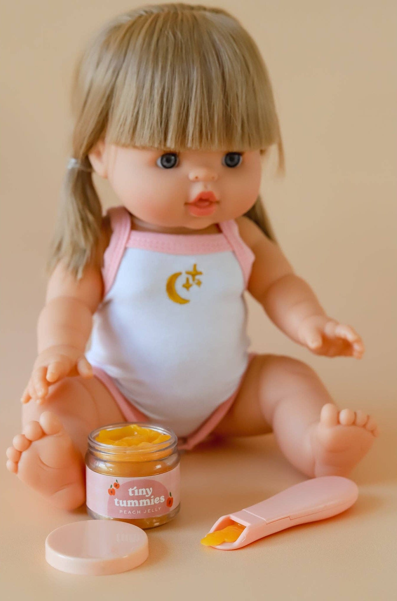 Doll wearing a white onesie with a pink trim, sitting next to a jar labeled 'tiny tummies' on a beige background.