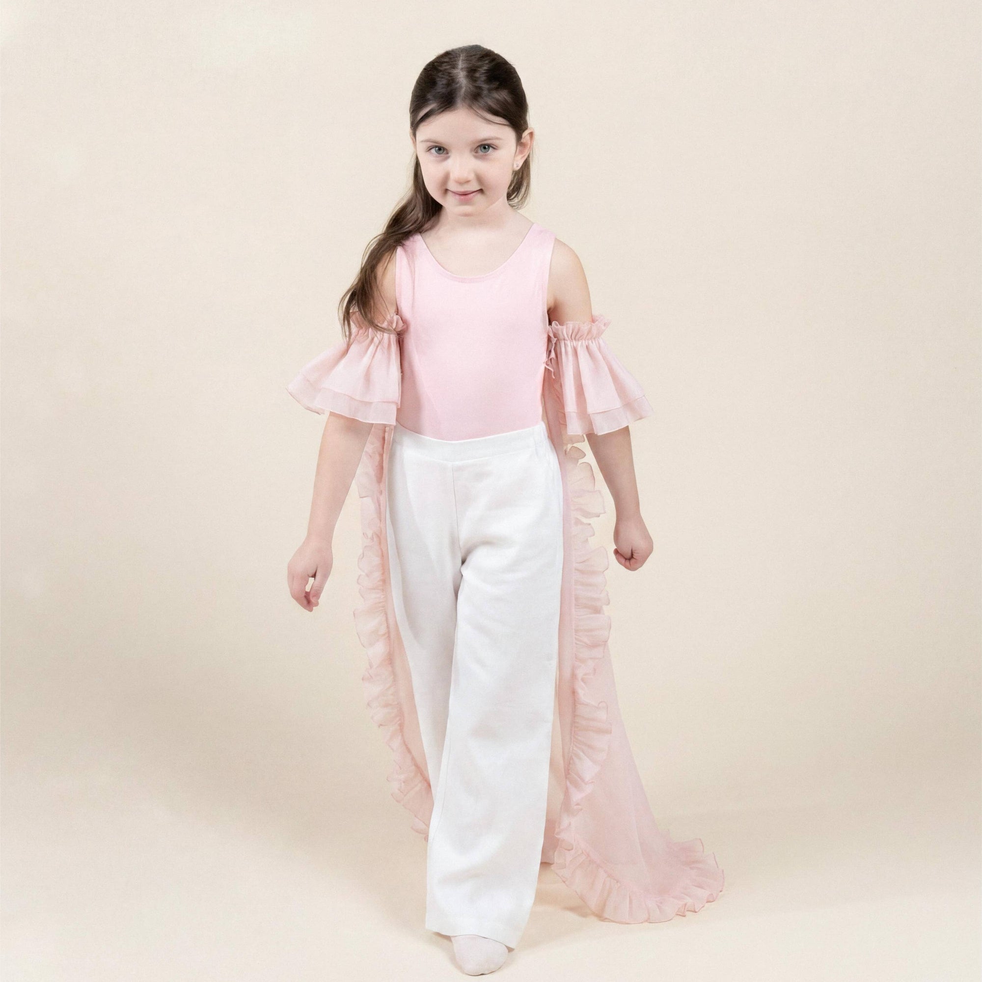 A young girl modeling a light pink cold-shoulder ruffle top and white wide-leg pants against a soft beige studio background.