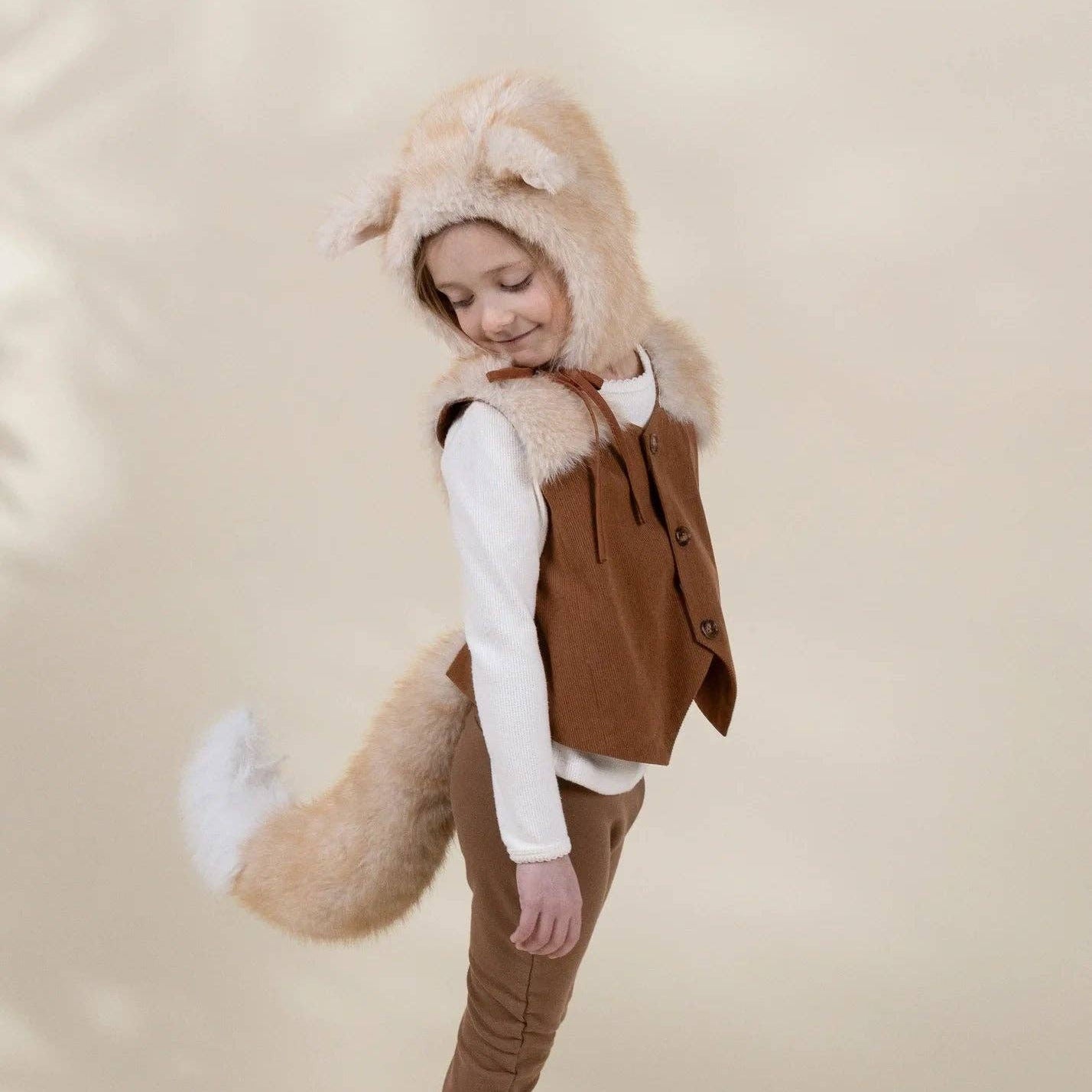 A child wearing a woodland animal costume, featuring a brown faux-fur vest, matching brown pants, a white long-sleeve shirt, and a hood with a fluffy beige tail and ears, standing against a plain beige background.